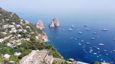 Havadan bakıldığında, Capri, İtalya açıklarında ikonik Faraglioni kaya oluşumları görülüyor. Dalgalı mavi denizi, yemyeşil yamaçların yanında binalarla dolduran sayısız tekne var..