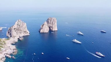 Havadan bakıldığında, Capri, İtalya açıklarında ikonik Faraglioni kaya oluşumları görülüyor. Dalgalı mavi denizi, yemyeşil yamaçların yanında binalarla dolduran sayısız tekne var..