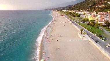 Bir hava manzarası, kıyı kasabası ve paralel giden bir karayolu ile Tyrhenian Denizi 'nin uzun kumlu ve turkuaz sularını yakalar. İnsanlar açık gökyüzünün altında güneşin tadını çıkarırlar..