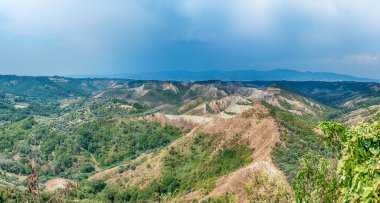 Açık renkli 'calanchi' çorak arazilerinin dramatik jeolojik oluşumlarını, yeşil ağaçlar ve kuru bitki örtüsüyle kaplı puslu mavi gökyüzünün altında görüntüler..