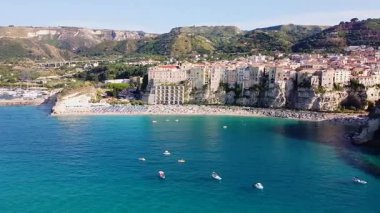 İtalya 'nın Tropea kentinin pitoresk rıhtımlarını gösteren hava manzarasında canlı bir plaj, yüksek beyaz kayalıklar ve turkuaz denize bakan tarihi binalar berrak bir gökyüzünün altında yer alıyor..