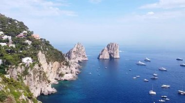 Capri kıyılarında ikonik Faraglioni kaya oluşumlarını gösteren bir hava görüntüsü var. Sayısız yat ve tekne açık bir günde koyu mavi Tiren Denizi 'ni noktalıyor..