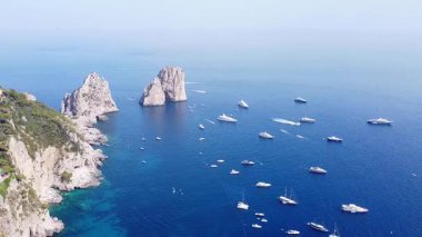 Capri kıyılarında ikonik Faraglioni kaya oluşumlarını gösteren bir hava görüntüsü var. Sayısız yat ve tekne açık bir günde koyu mavi Tiren Denizi 'ni noktalıyor..