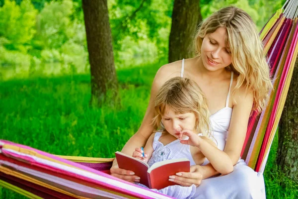自然公園でハンモックで本を読んでいる子供を持つ幸せな母親 — ストック写真