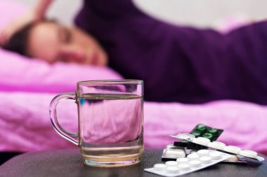 Bir kadın kendini hasta hisseder ve yanında tablet şeklinde bir bardak su ve ilaç vardır..