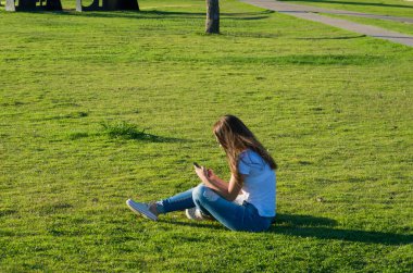 Genç bir kadın çimlerin üzerinde oturuyor. Yeni Orla do Guaiba 'da akıllı telefon kullanıyor. Güzel bir günbatımı gününde Porto Alegre' de.