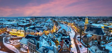 Kışın gün batımında karla kaplı tarihi bir deniz kasabasının panoramik günbatımı manzarası. Sıcak akşam ışığı, sakin ve şenlikli bir kış atmosferi yaratıyor. ve limandaki eski gemileri aydınlattı. .