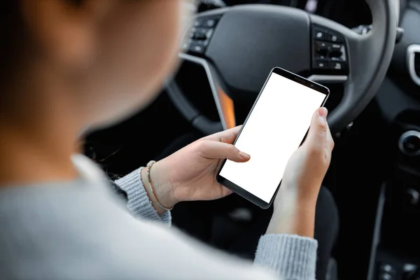 Young driver using smartphon in the car while driving or parking. Mockup of a mobile phone with isolated white screen in the hands of a woman.