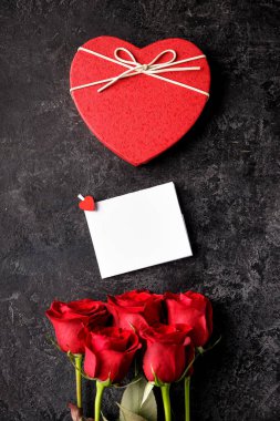 Kırmızı şömine şeklinde hediye kutusu, güller, siyah bir grunge masasında boş bir mesaj kartı. Sevgililer Günü arkaplanı.