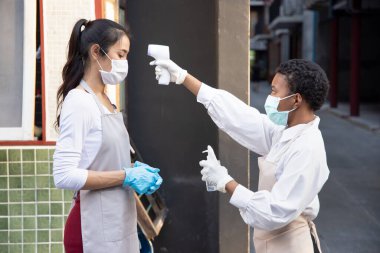 Afrikalı siyahi kadın perakende satış elemanları veya bakkal bakkalı taşınabilir termo tarama cihazı ile Asyalı iş arkadaşının ateşini kontrol ediyor; yeni normal iş uygulaması kavramı