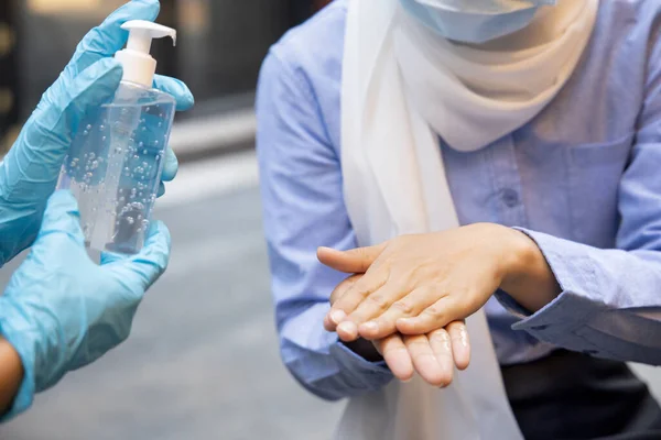 Asian woman employee or shop keeper wearing rubber glove while dispensing alcohol gel, hand sanitizer to Muslim woman customer; concept of new normal business practice with inclusive people diversity