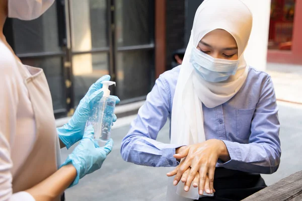 Asian woman employee or shop keeper wearing rubber glove while dispensing alcohol gel, hand sanitizer to Muslim woman customer; concept of new normal business practice with inclusive people diversity