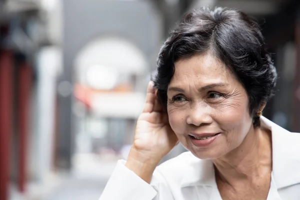 Asian senior woman listening to something; portrait of happy old asian female elderly with hearing ear, concept of rumor, gossip, good news, hearing loss, hard of hearing with correction aid device