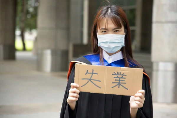 Text in traditional Chinese or Japanese Kanji reads Unemployed, banner carried by college graduate student woman wearing face mask, concept of unemployment, job cut after COVID 19 economic recession