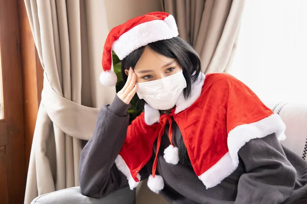 Unhappy Xmas woman in Santa clause suit having headache wearing face mask due to coronavirus and COVID-19 outbreak in Christmas period, concept of Covid Christmas or new normal social distancing Xmas