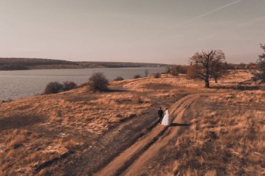 Gelin ve damat gelinlikle sonbahar kırmızı tarlasında nehir kenarında yürüyorlar.