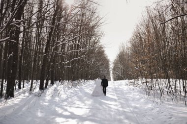 Yeni evli bir çift kışın ormanın ortasında, karlı bir yolda yürüyor.