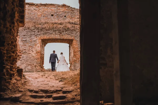 Bruid en bruidegom houden elkaars hand vast tegen de achtergrond van bakstenen ruïnes — Stockfoto
