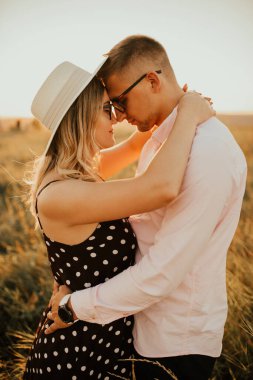 Şapkalı bir kadınla bir adam çayırdaki uzun otların arasında sarılıp öpüşüyor.