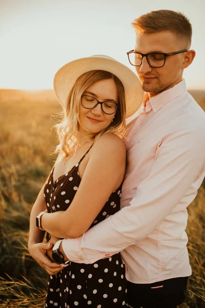 A man with a woman in a hat hug and kiss in the tall grass in the meadow — Stock Photo, Image