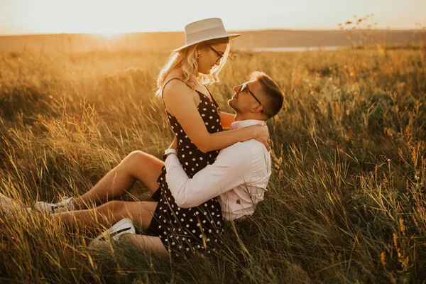 Dívka v klobouku sedí v rukou chlap na zemi uprostřed vysoké trávy na louce — Stock fotografie