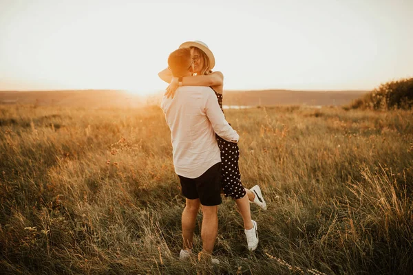 A man with a woman in a hat hug and kiss in the tall grass in the meadow — Stock Photo, Image