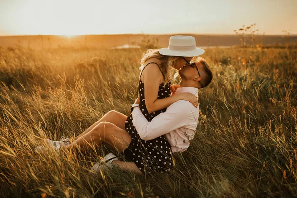 Una chica con sombrero se sienta en las manos chico en el suelo en medio de la hierba alta en el prado —  Fotos de Stock