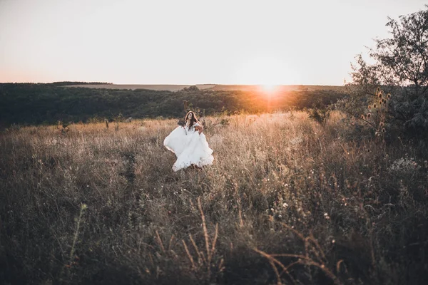 Braut im weißen Hochzeitskleid spaziert im Sommer bei Sonnenuntergang über die Wiese — Stockfoto