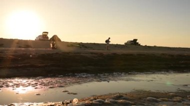 Uzun saçlı bikinili bir kadın gün batımına karşı deniz kıyısında oturuyor.