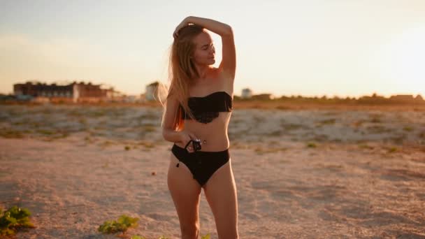 Mujer rubia en un traje de baño bikini negro alisar los pies de pelo largo en la playa puesta el sol — Vídeos de Stock