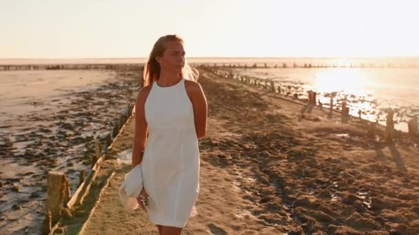 Mujer rubia bronceada en vestido blanco se para al atardecer en la playa — Vídeos de Stock
