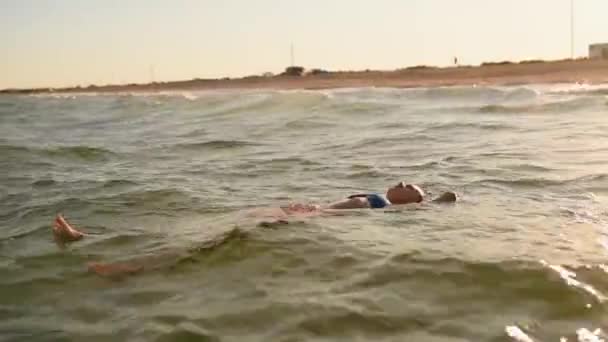 Młoda sexy kobieta pływa w morze na dużych falach w bikini strój kąpielowy o zachodzie słońca — Wideo stockowe