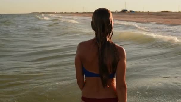 Giovane donna sexy che cammina in mare sulle grandi onde in bikini costumi da bagno al tramonto — Video Stock
