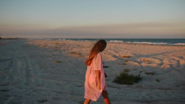 Mujer sexy en un traje de baño de bikini rosa y capa de playa camina a lo largo de la orilla del mar de arena al atardecer en el viento — Vídeo de stock