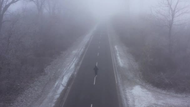 冬日里，穿着运动服的男子在多雾的天气里沿着被霜冻树环绕的道路奔跑。来自无人机的4K航拍视频 — 图库视频影像