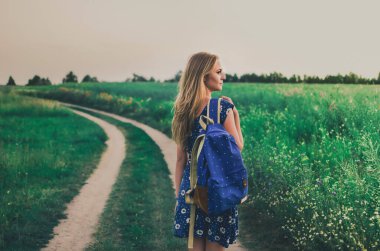 Kadın yeşil bir tarlanın ortasında bir elbise ve sırt çantası içinde yürüme yolunda duruyor.