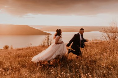 Beyaz elbiseli ve takım elbiseli, sevgi dolu bir çift evleniyor. Nehrin yukarısındaki dağda, uzun çimlerin üzerinde mutlu mesut gülümsüyor. gün batımı.