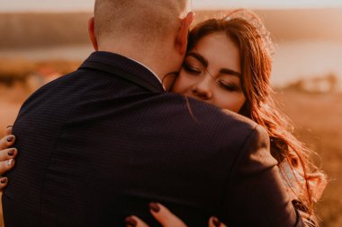 Beyaz elbiseli ve takım elbiseli, sevgi dolu bir çift. Nehrin yukarısındaki dağda uzun çimenlerin üzerinde öpüşen uzun boylu bir çift. Saç stili saç stili üzerinde gelin aksesuarları.