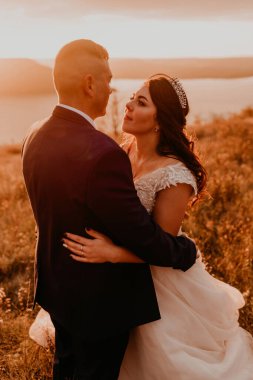 Beyaz elbiseli ve takım elbiseli, sevgi dolu bir çift. Nehrin yukarısındaki dağda uzun çimenlerin üzerinde öpüşen uzun boylu bir çift. Saç stili saç stili üzerinde gelin aksesuarları.