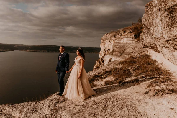 loving couple wedding newlyweds in white dress and suit walk in summer on mountain above river. sunset and sunrise. man and woman on rocks above cliff