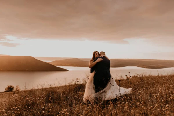 loving couple wedding newlyweds in white dress and suit hug kissing whirl on tall grass in summer field on mountain above the river. sunset.