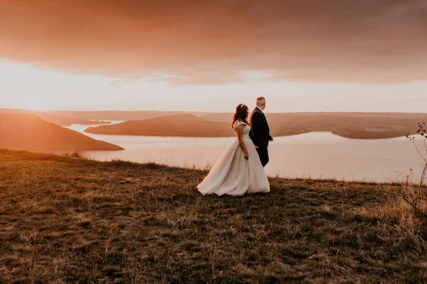 A loving couple wedding newlyweds walk in tsummer in the fall on mountain on cliff above the river. sunset. bride in a white dress with crown on her head. style fashionable women hairstyle makeup