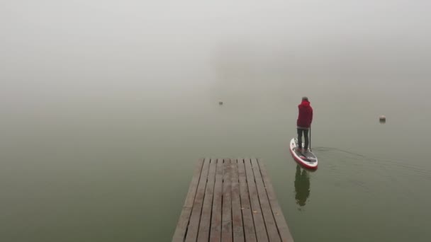 Een man staat op een waterschap drijft vanaf een houten pier langs een gladde kalme rivier in dikke mist — Stockvideo