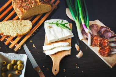 Tahta bir tabakta dilimlenmiş peynir taze pişmiş ekmekle taze soğan, füme jambon, sosis, siyah ahşap bir yemek masasında zeytin.