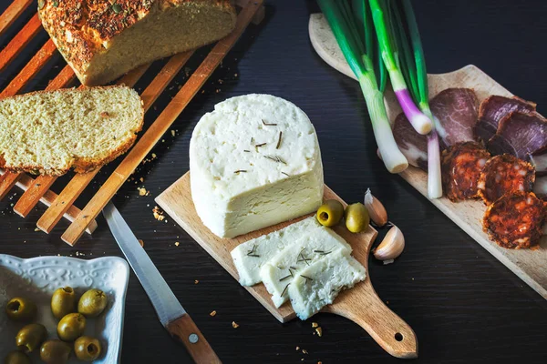 Tahta bir tabakta keçi peyniri dilimlenmiş taze pişmiş ekmekle taze soğan, füme jambon, sosis, siyah ahşap bir yemek masasında zeytin.