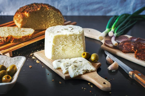 Tahta bir tabakta keçi peyniri dilimlenmiş taze pişmiş ekmekle taze soğan, füme jambon, sosis, siyah ahşap bir yemek masasında zeytin.