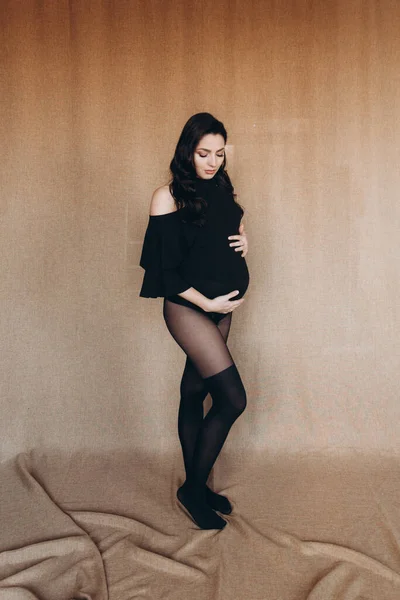 Pregnancy photo session in a photo studio with a fabric background in a black bodysuit and tights