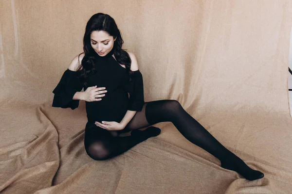 Pregnancy photo session in a photo studio with a fabric background in a black bodysuit and tights