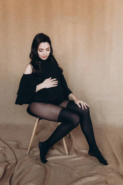 Pregnancy photo session in a photo studio with a fabric background in a black bodysuit and tights