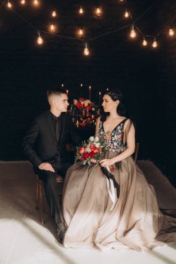 Siyah elbiseli güzel bir çiftin düğünü, Marsala renginde bir buket, mumlarla dolu bir şenlik masasında ve siyah bir duvara yaslanmış bir pasta.